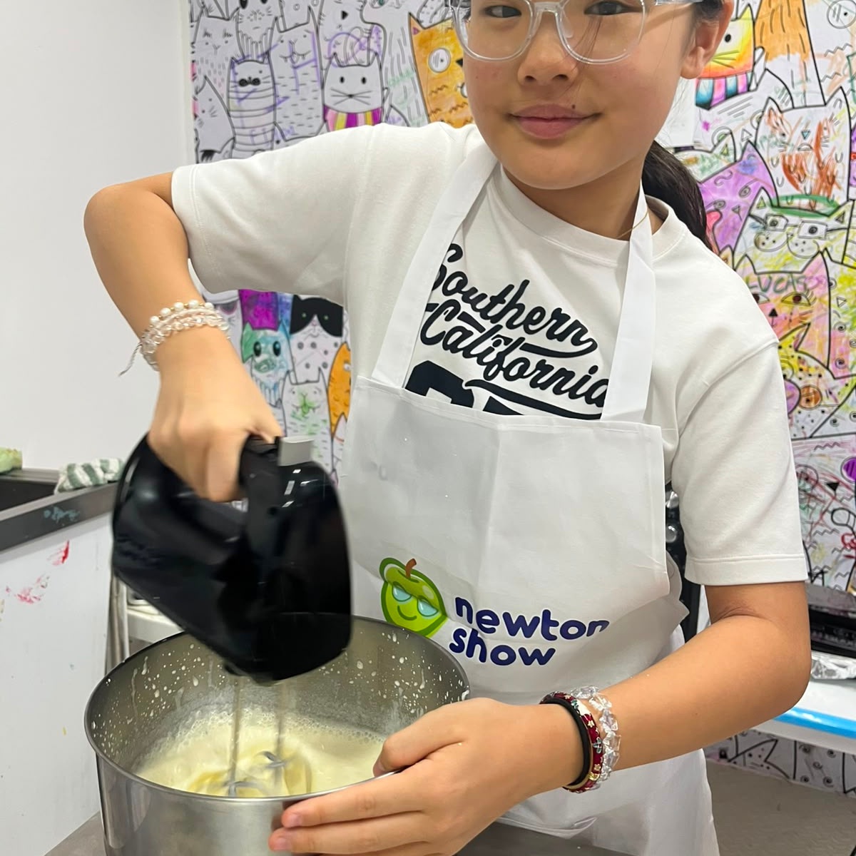 Girl in a Newtonshow apron using a hand mixer during a camp activity